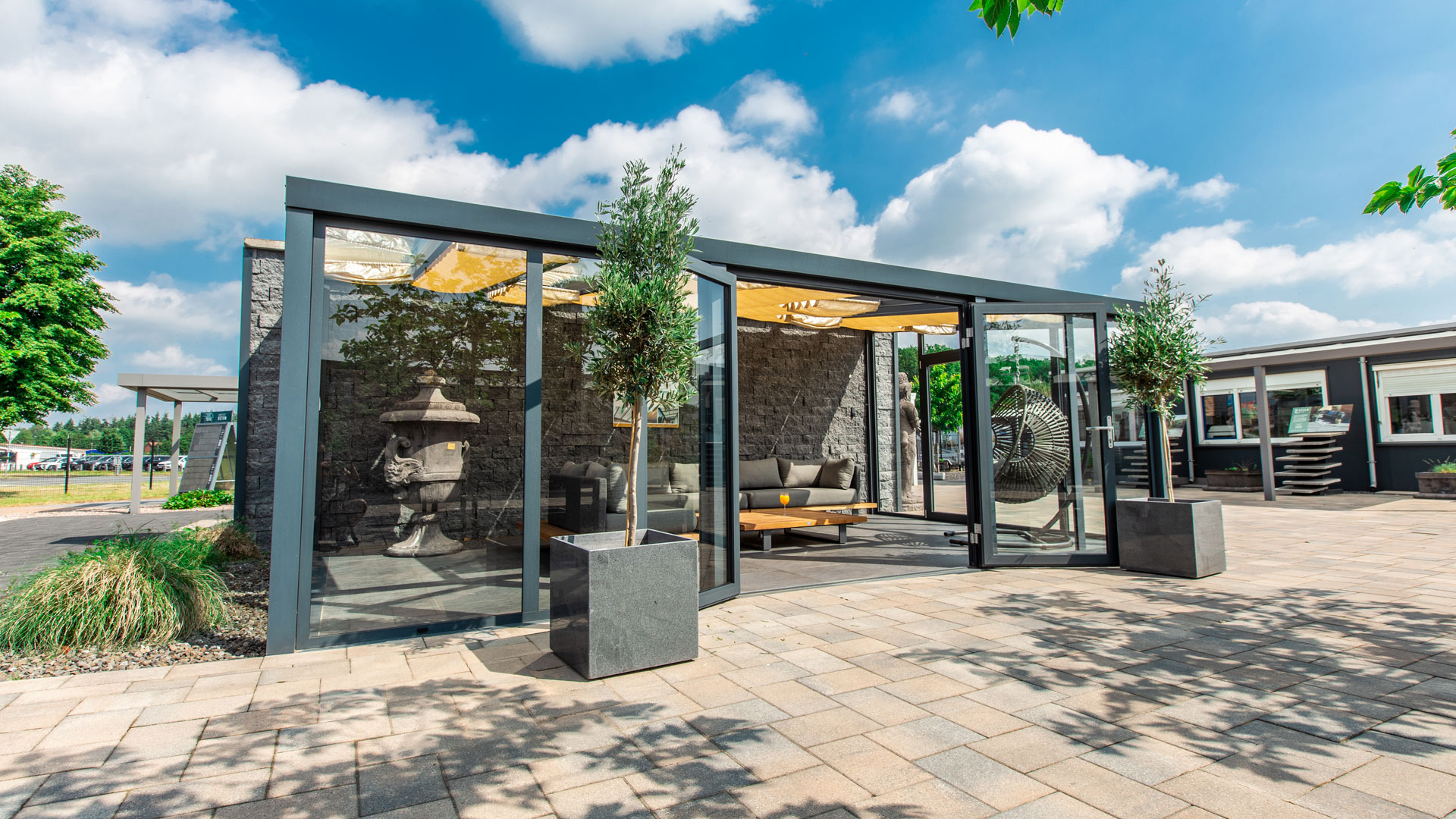 Moderner Wintergarten mit Glaswänden, grauen Möbeln und Bäumen. Himmel blau mit weißen Wolken.