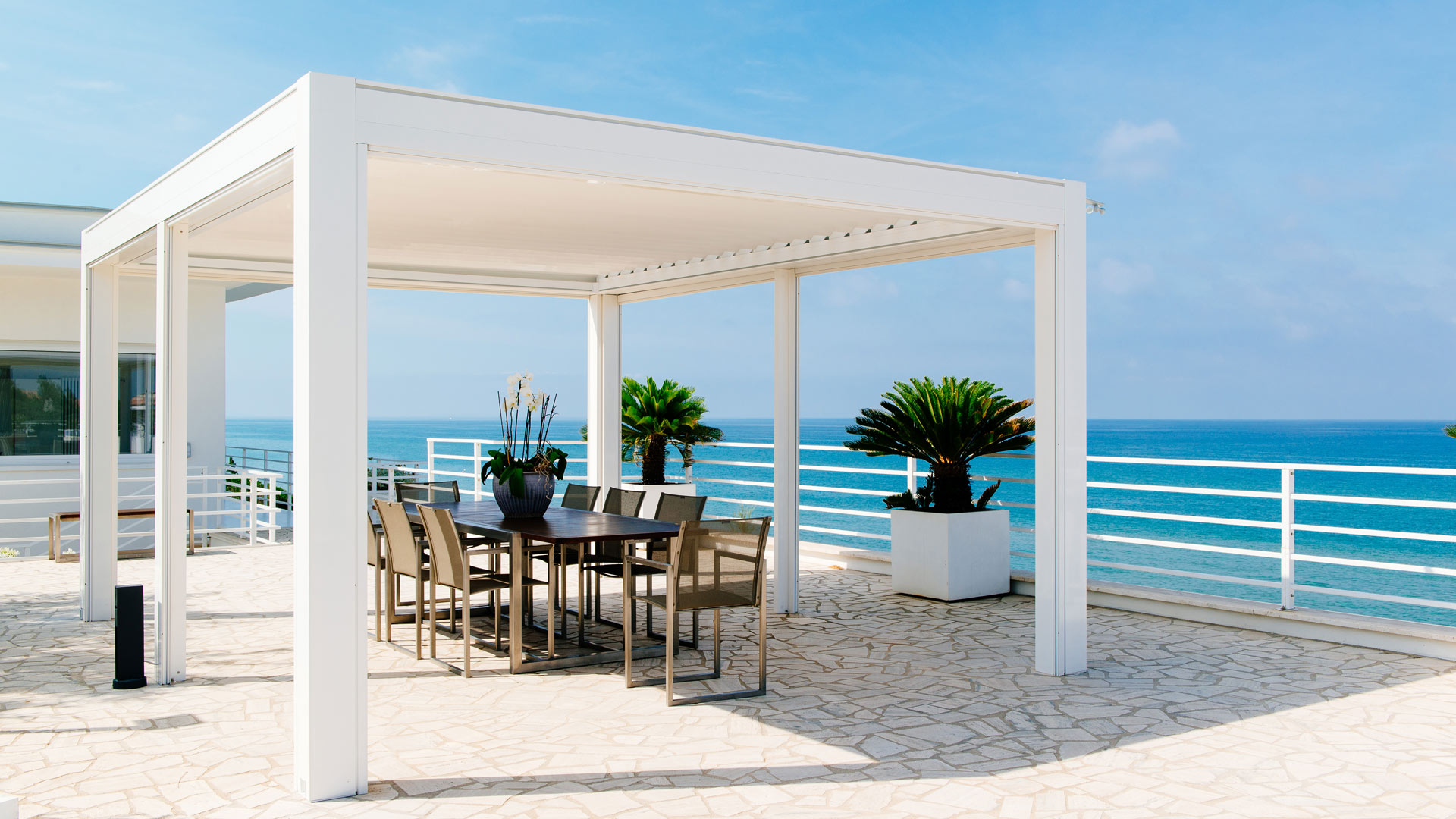 Ein weißer Pavillon auf einer Terrasse mit Meerblick; Tisch, Stühle, und Pflanzen im Vordergrund.
