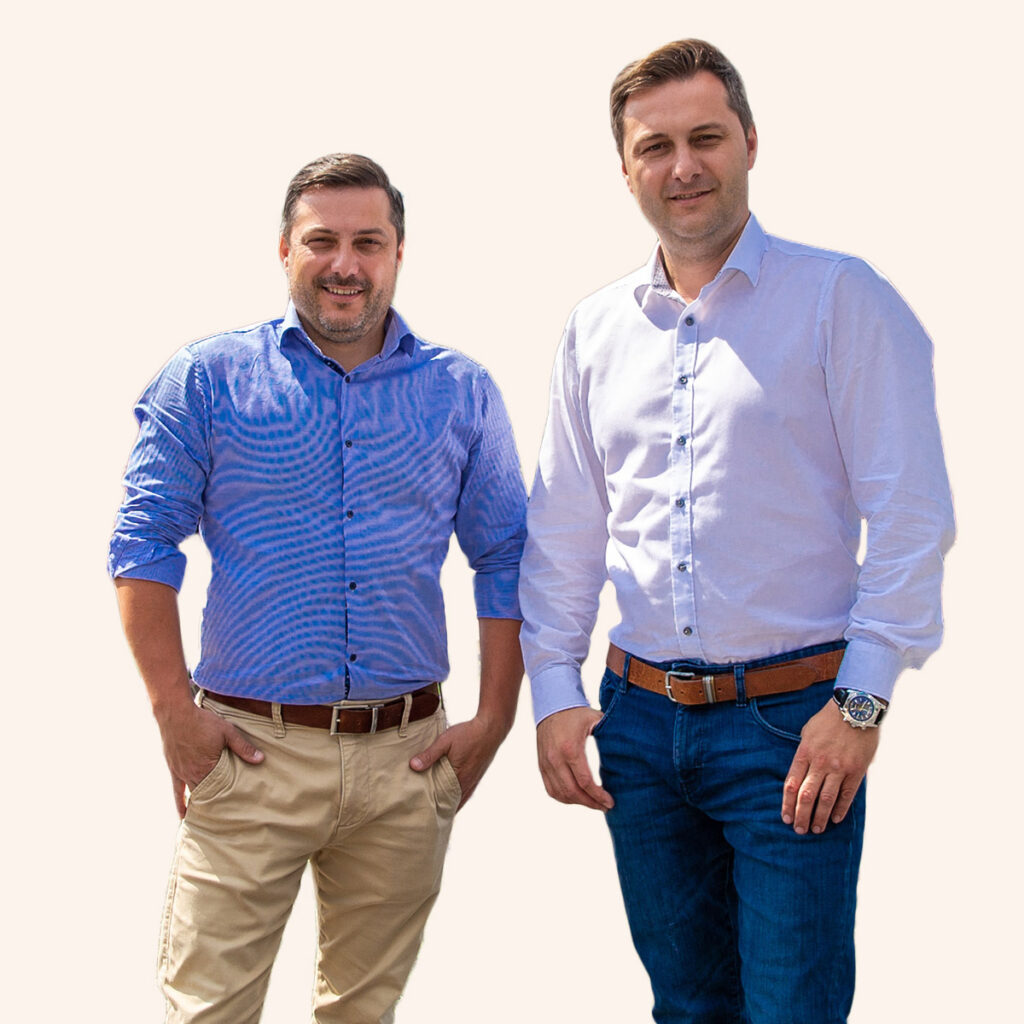 Zwei Männer lächeln, tragen helle Hemden, blaue Jeans; einer hat beige Hosen, beide mit Uhren.